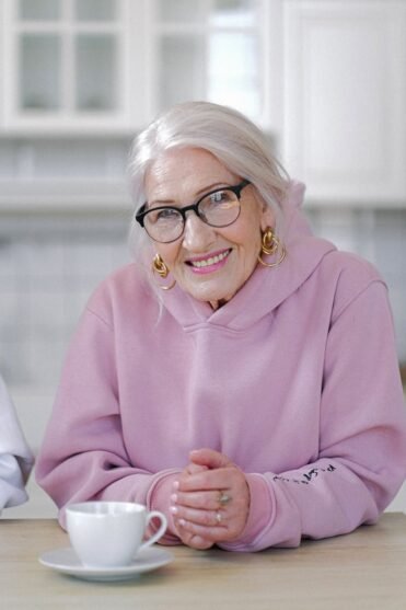 Smiling aged woman in hoodie sitting at table in light apartment